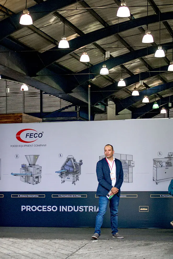 Muro de Stand con productos ilustrados necesarios para un proceso industrial de panadería. Alfrente dos personas caminan para un lado y otra en sentido contrario.